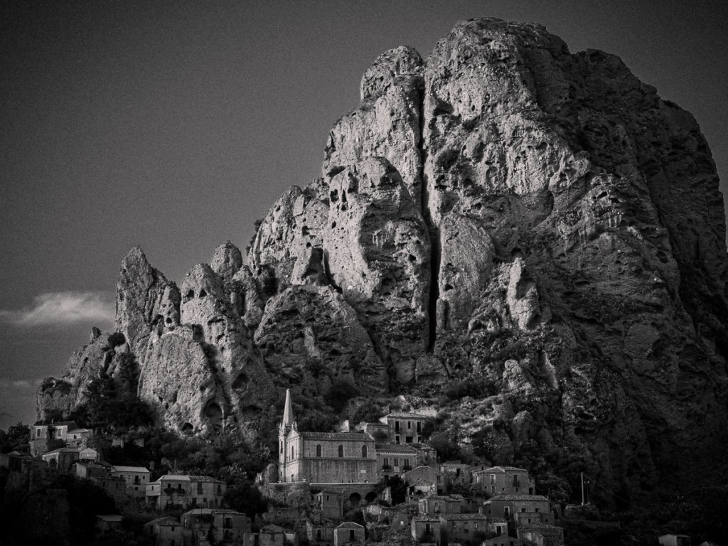 A black and white photograph of the ghost town of Pentedattilo, Calabria, showing stone buildings and a church nestled beneath a towering, rugged cliff shaped like a petrified hand.