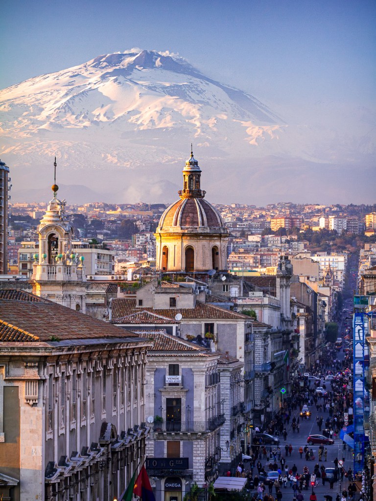 View from the terrace of the Diocesan Museum in Catania: the city’s historic center with domed churches and lively streets in the foreground, and snow-capped Mount Etna rising in the background.