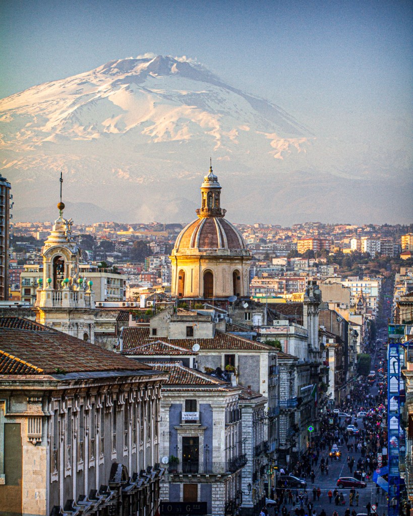 View from the terrace of the Diocesan Museum in Catania: the city’s historic center with domed churches and lively streets in the foreground, and snow-capped Mount Etna rising in the background.