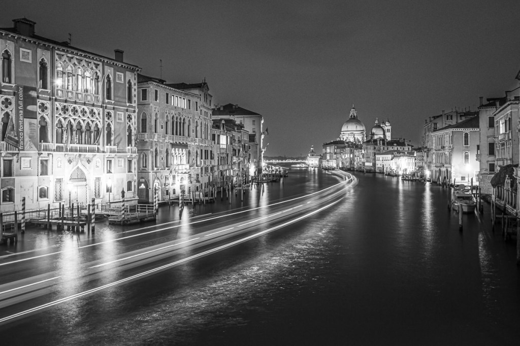 Long-exposure black and white photograph of Venice’s Grand Canal at night, featuring light trails from passing boats, historic buildings with ornate facades, and the distant dome of the Basilica di Santa Maria della Salute.