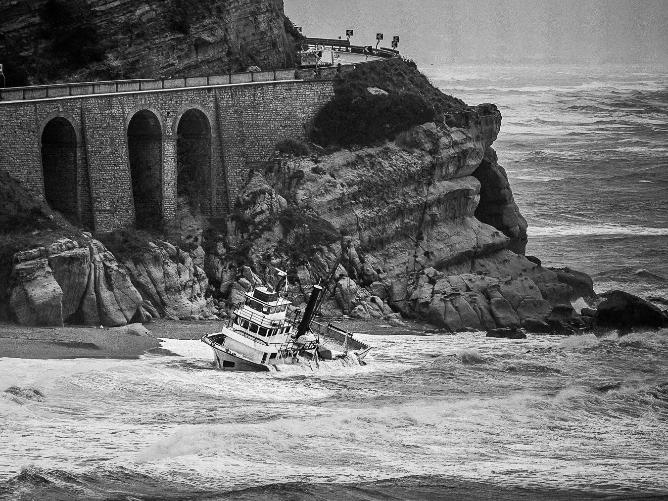 Black and white photo of a fishing boat wreck leaning in the waves beneath stratified cliffs and a road, Capo Bruzzano, Calabria