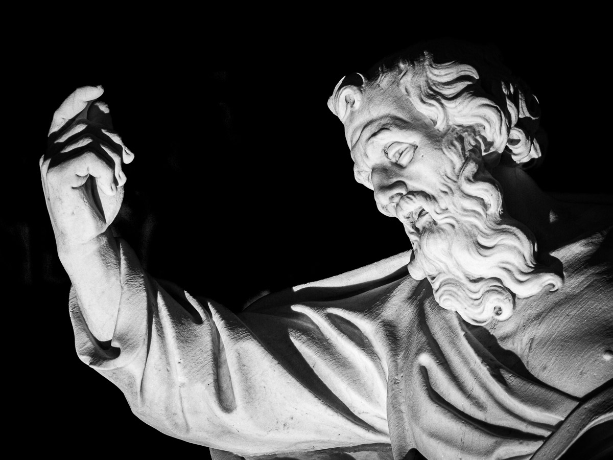 Close-up of the marble statue of Saint Paul in front of the Cathedral of Ortygia, dramatically lit against a black background, highlighting his expressive gesture and detailed features.