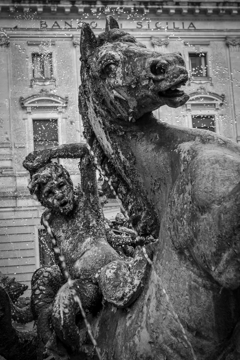 A dramatic black-and-white detail of the Diana Fountain in Ortygia, featuring a rearing horse and mythological figure amid dynamic sprays of water, with the historic Banco di Sicilia building in the background.