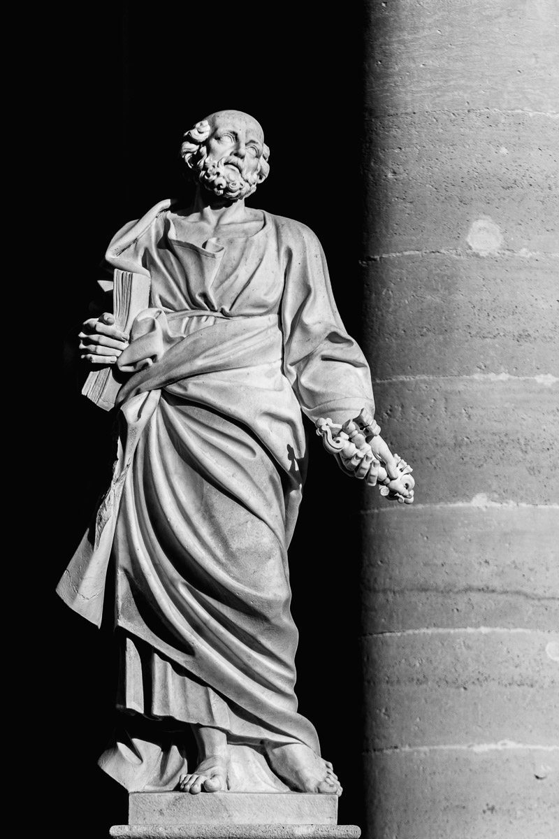 Marble statue of Saint Peter in front of a columned façade in Ortygia, holding a large key and a book, dramatically lit against a dark background to emphasize the intricate carving and symbolic gesture.