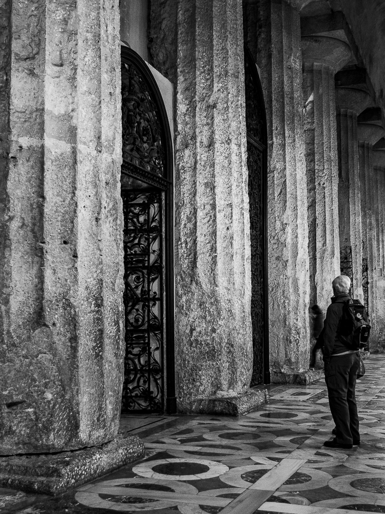 Black and white photo of a person standing before massive stone columns and wrought iron gates at the Cathedral of Ortygia in Siracusa, Sicily.