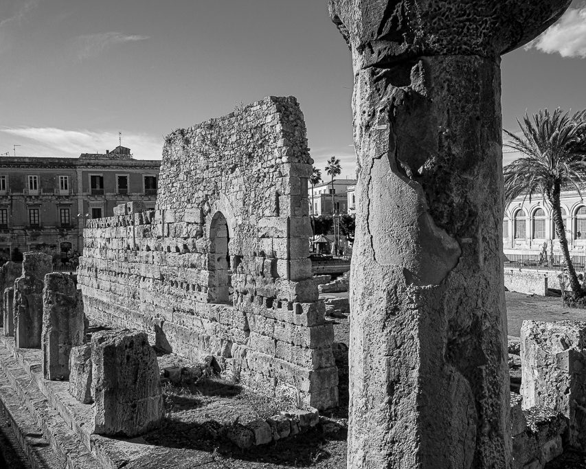 Black and white photo of the ruins of the Apollo Temple in Ortygia, Siracusa, showing weathered stone columns and arches with modern buildings and palm trees in the background.