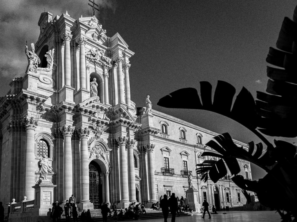 Black-and-white photograph of the Duomo of Ortygia in Siracusa, Sicily, showing its ornate baroque façade with columns, statues, and intricate carvings. People stroll and sit in the sunlit square, while large silhouetted tropical leaves frame the right side of the image.