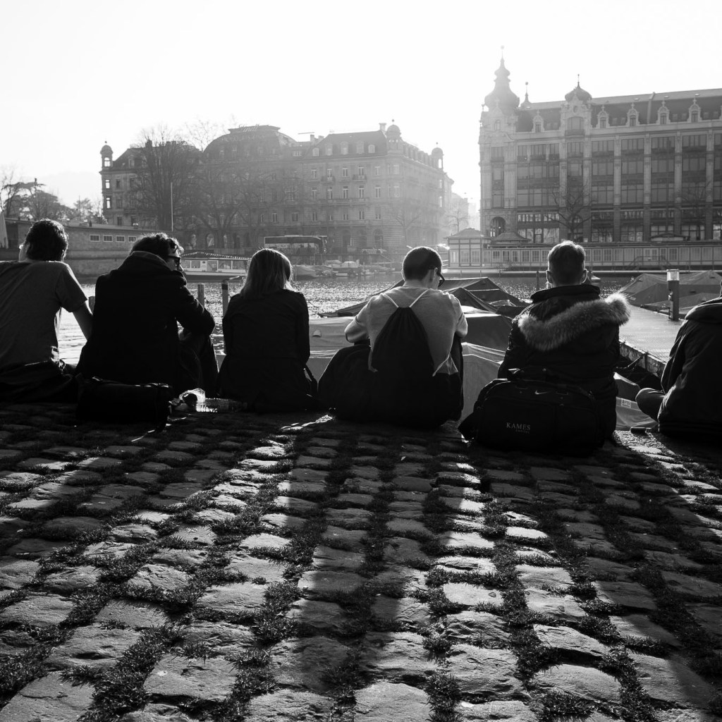 People sitting by Limmat river at sunset watching Zurich cityscape, black and white street photography