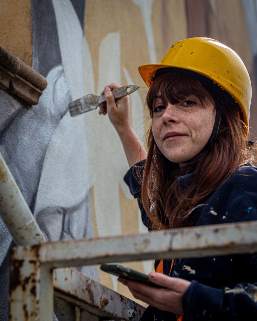 Roberta Fiorito, known as Rob_arkt, stands on scaffolding while painting a large mural on the weathered façade of a public housing block in Brancaleone, Calabria. The wall shows peeling plaster and faded colors, now being transformed by her brush.