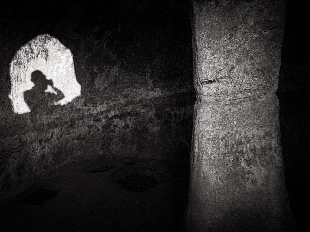 Sunlight filters through a small opening, illuminating a carved stone pillar known as the Tree of Life inside a cave-church in the abandoned hilltop village of Brancaleone Superiore, Calabria.