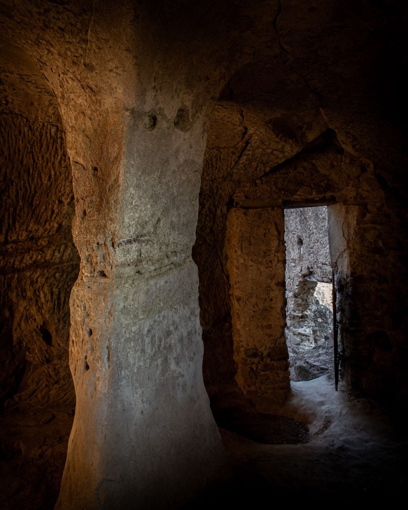 Interior of the Armenian cave church in Brancaleone Superiore, Calabria, with rough stone walls, a central pillar, and natural light streaming through the doorway into the dim chamber.