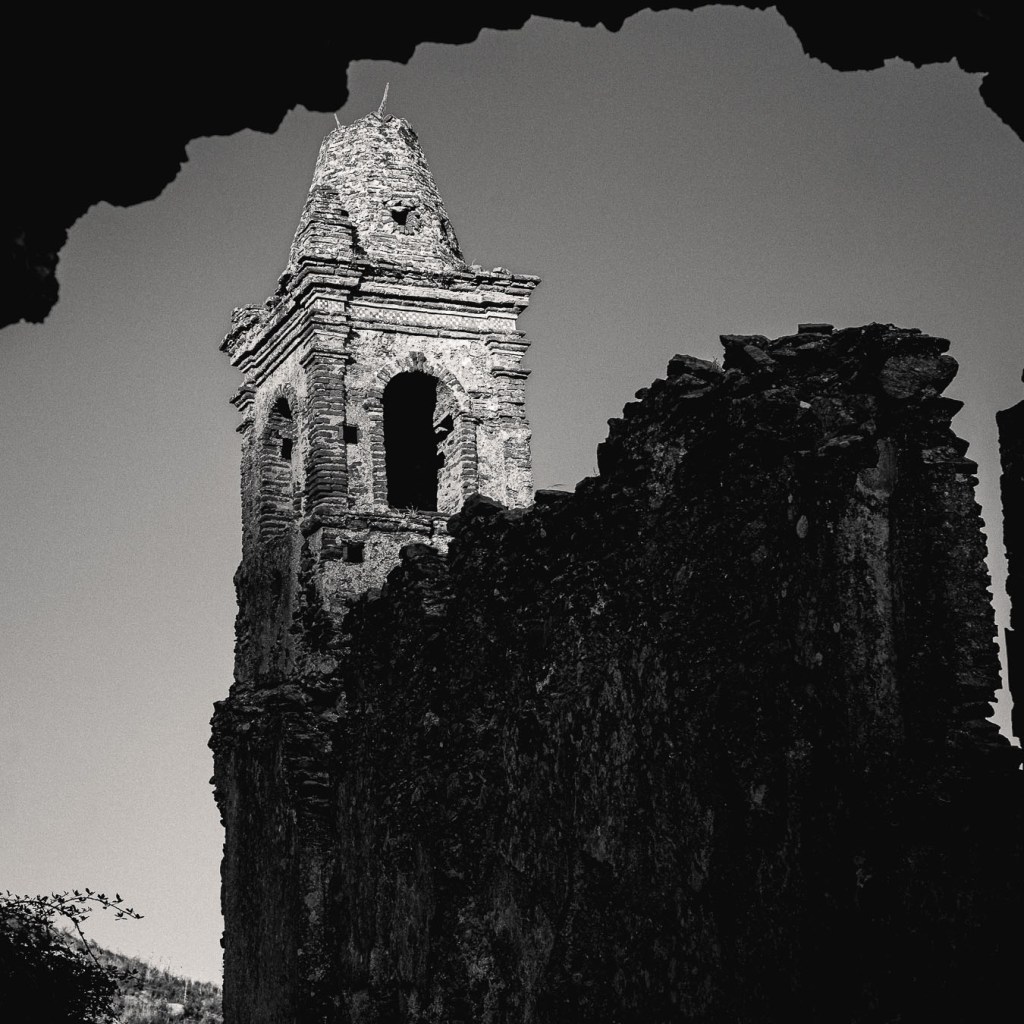 Black and white photograph of the ruined bell tower of Santa Maria dell’Alica framed by jagged stone, with clear sky behind; weathered arches and crumbling walls evoke centuries of abandonment.