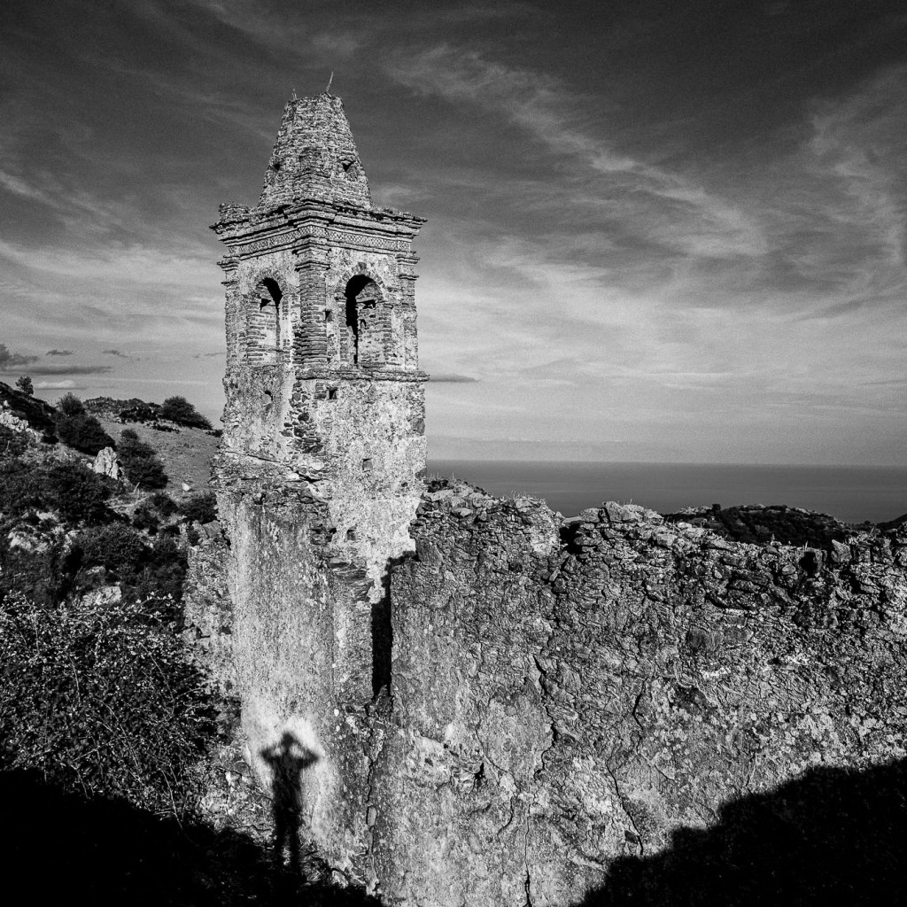 Black and white photograph of the ruined bell tower of Santa Maria dell’Alica framed by jagged stone, with clear sky behind; weathered arches and crumbling walls evoke centuries of abandonment.