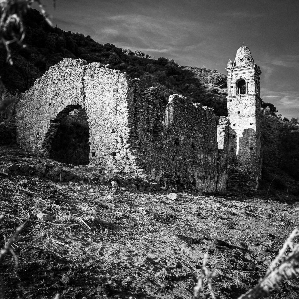 Black‑and‑white photograph of the stone ruins of Santa Maria dell’Alìca in Pietrapennata, Calabria, showing a tall, narrow bell tower with a pointed roof and a partially collapsed archway, set on a rugged hillside with sparse vegetation.