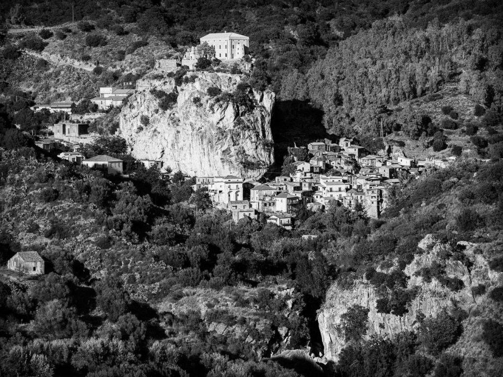 Black and white photograph of Palizzi Superiore, a hillside village in Calabria, Italy, showing stone buildings clinging to a rocky slope beneath a dramatic cliffside mansion, surrounded by wild vegetation and rugged terrain.