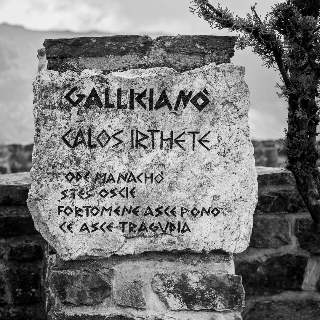 Stone plaque in Gallicianò, Condofuri, Calabria, engraved with stylized Greek text, mounted on a stone wall in a rural mountain setting with tree and natural background.