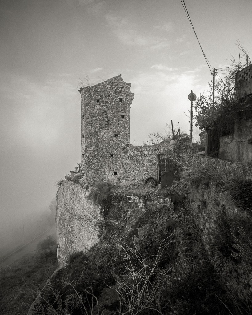 An ancient stone house stands on a rocky outcrop, partially ruined, with overgrown vegetation and power lines visible against a misty sky. Ferruzzano Superiore, Calabria.