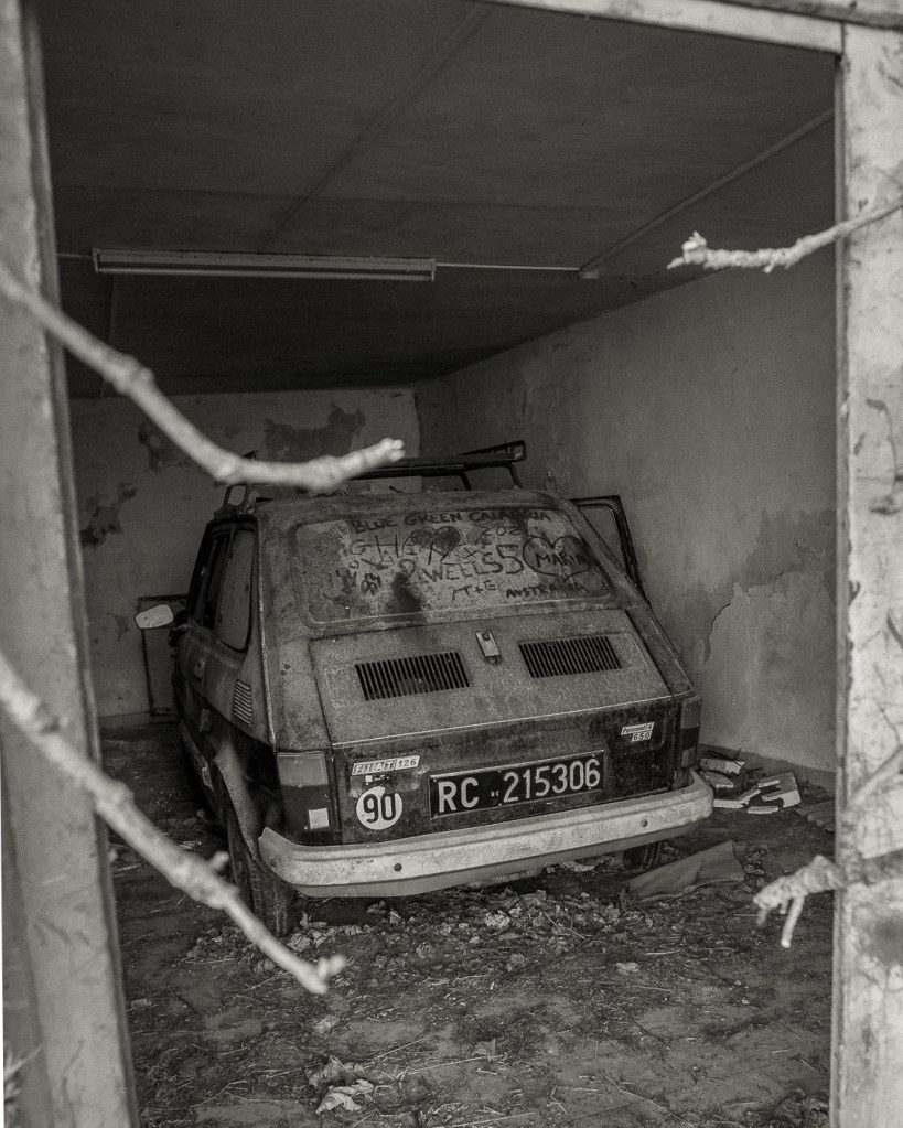An abandoned Fiat 126 with license plate RC 215306 sits in a garage, covered in dust, with handwritten text visible on the rear windshield. Ferruzzano Superiore, Calabria