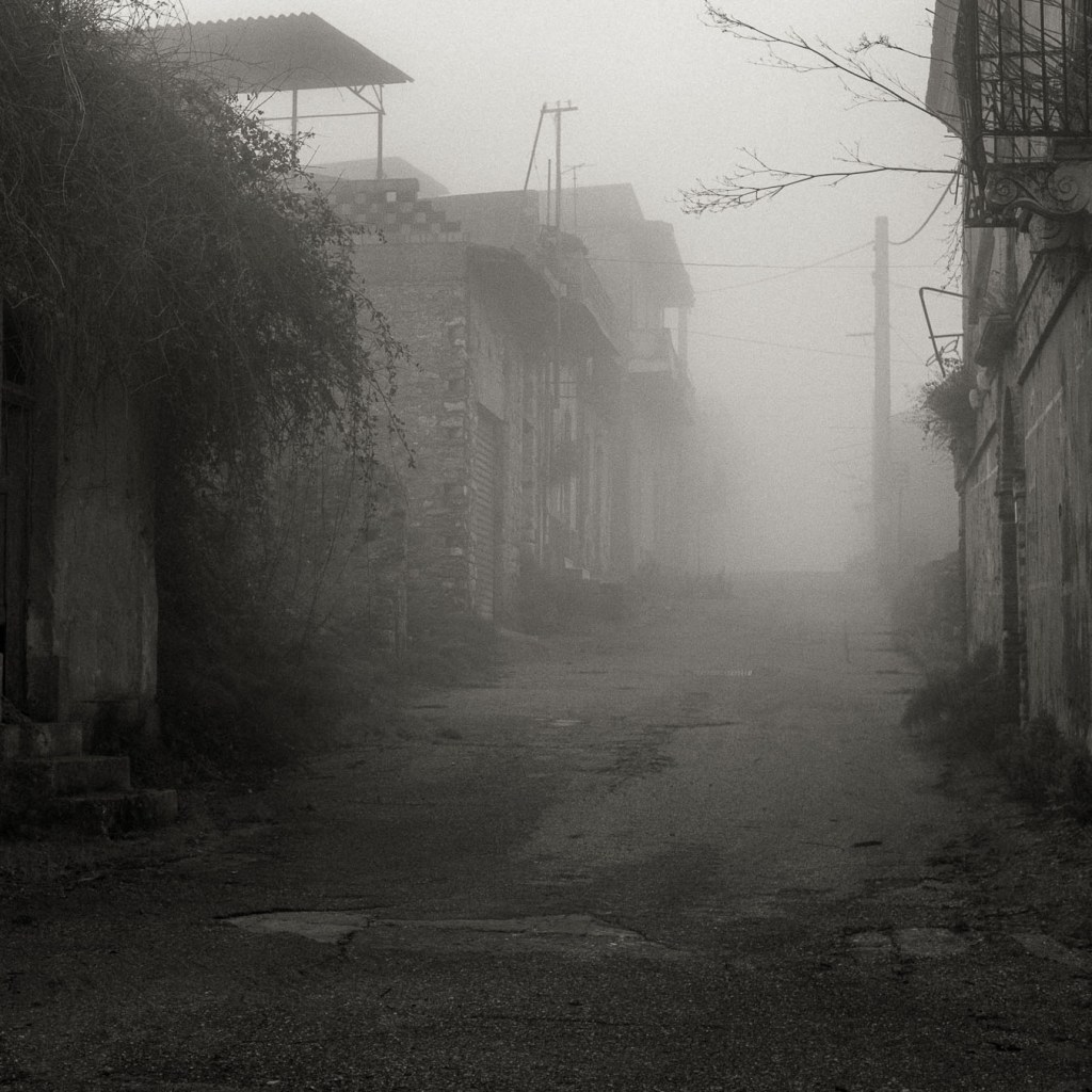 A foggy street in an abandoned village disappears into thick mist, with old stone buildings barely visible on both sides. Ferruzzano, Calabria