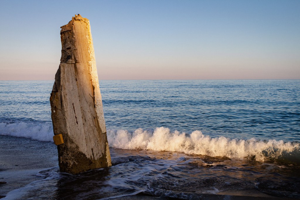 A damaged concrete pillar standing in shallow sea water, waves breaking around its base, at sunset. Ionian Coast, Brancaleone, Calabria, Italy.