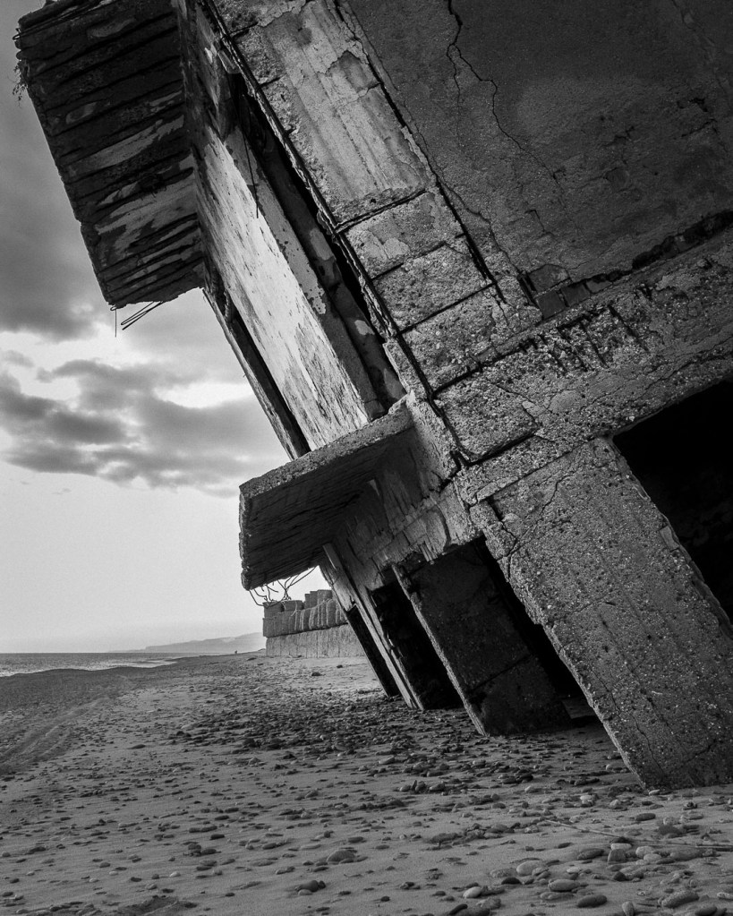Large, leaning concrete structure in ruins on the beach near Bova Marina; weathered surfaces with deep cracks and exposed rebar, with a calm sea and cloudy sky in the background.