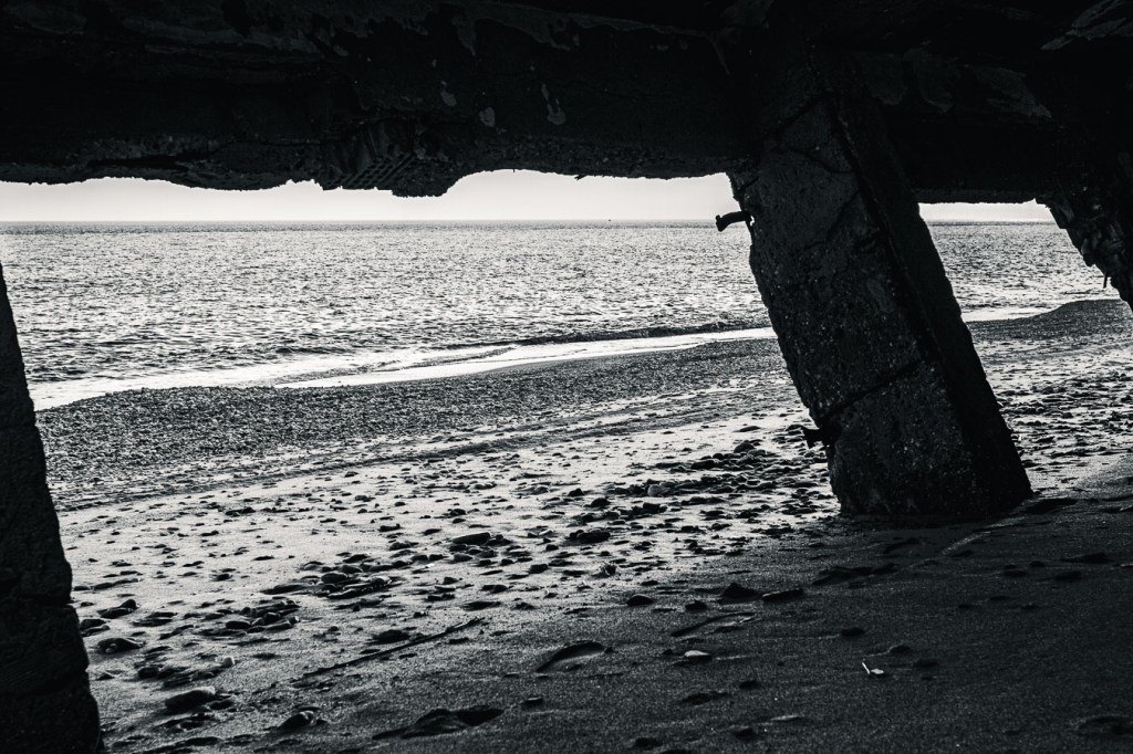 View from inside a collapsed concrete building on the beach near Bova Marina; broken beams and tilted walls frame the calm sea and distant horizon under an overcast sky.