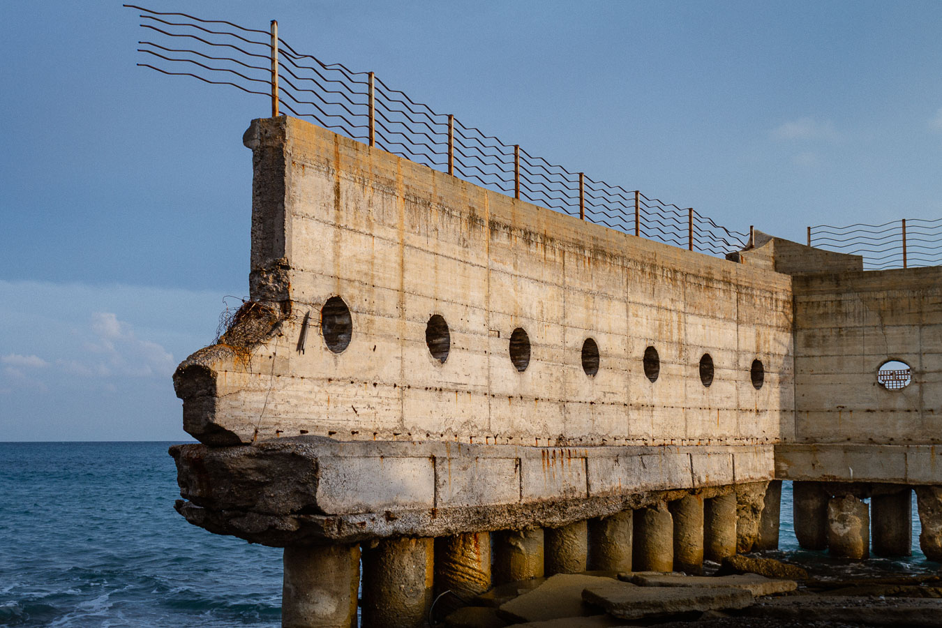 A large abandoned concrete structure on the sea, supported by cylindrical pillars, with a row of circular openings along its facade and corrugated metal rods along the top. Ionian Coast, Brancaleone, Calabria, Italy.