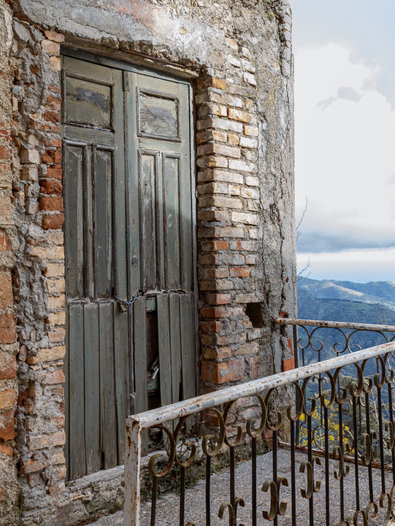 Weathered green wooden door set in a crumbling brick wall, mountain valley visible beyond a metal railing, Bova, Calabria