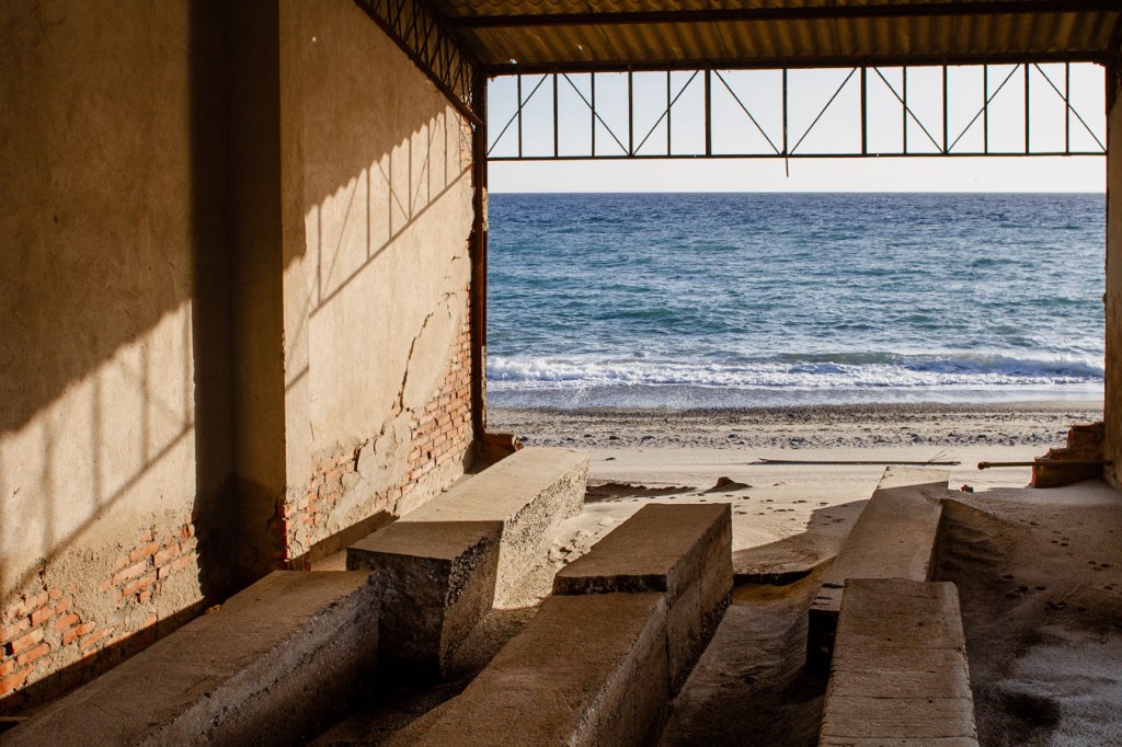 Interior of a ruined coastal structure open to the sea, with concrete benches, exposed brick walls, and a metal-framed roof casting shadows. Ionian Coast, Bova Marina, Calabria, Italy.