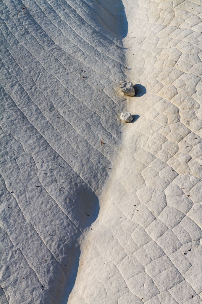 Aerial view of clay surface divided between shadow and light, two textures in contrast: parallel striations on the left, polygonal cracking on the right. Two small stones at the boundary. Calanchi di Palizzi, Calabria.