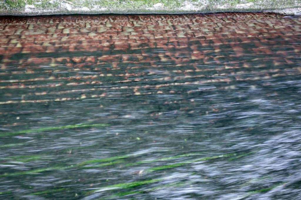 Canal surface seen from above: a brick bed visible through running water, green algae drawn out by the current.