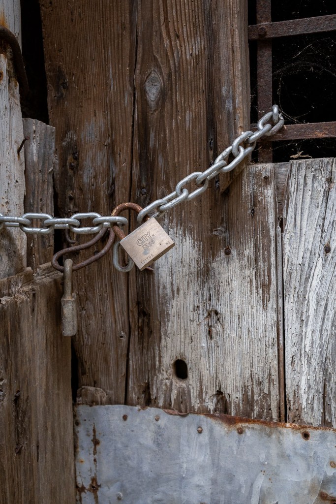 Chain and brass padlock across weathered wooden doors, next to an older rusted hook, with a barred window above right and a metal sheet below. Ferruzzano Superiore, Calabria.