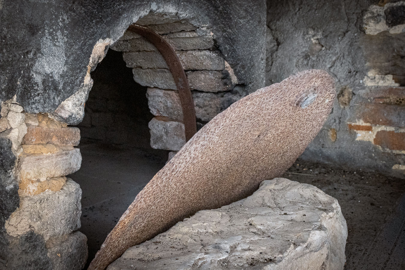 Broken opening in a dark grey plastered wall, revealing a brick-lined arch with an iron band, and a large round disc leaning against a block of pale stone. Ferruzzano Superiore, Calabria.