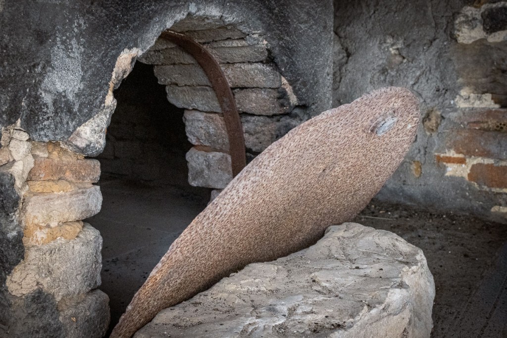 Broken opening in a dark grey plastered wall, revealing a brick-lined arch with an iron band, and a large round disc leaning against a block of pale stone. Ferruzzano Superiore, Calabria.